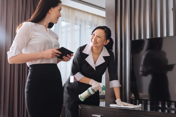 Hotel service. Joyful nice positive businesswoman standing near a hotel maid and talking to her while holding a tablet